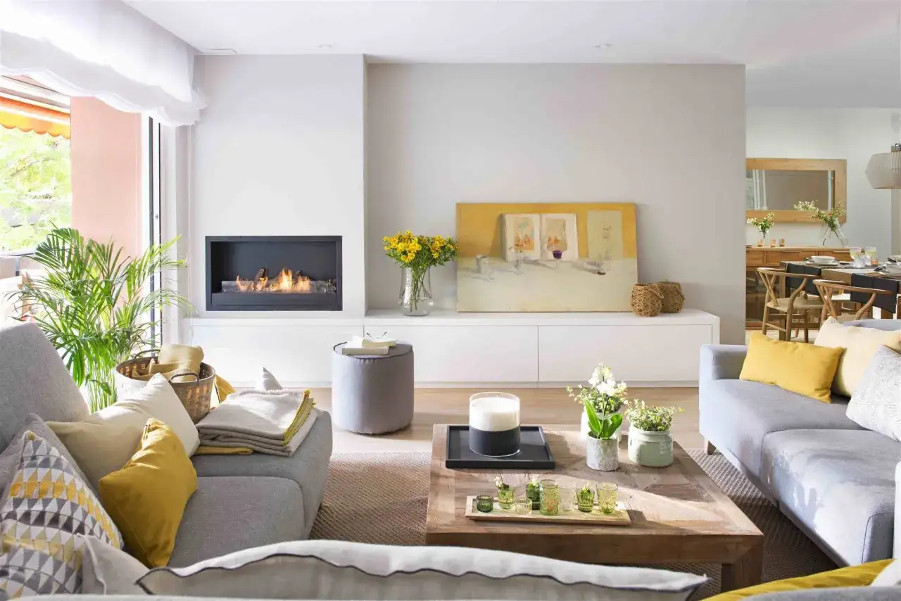 Elegante sal&oacute;n moderno donde se combina color gris en sof&aacute;s y puf con toques amarillos en cojines y arte. Chimenea encendida y plantas verdes aportan calidez.