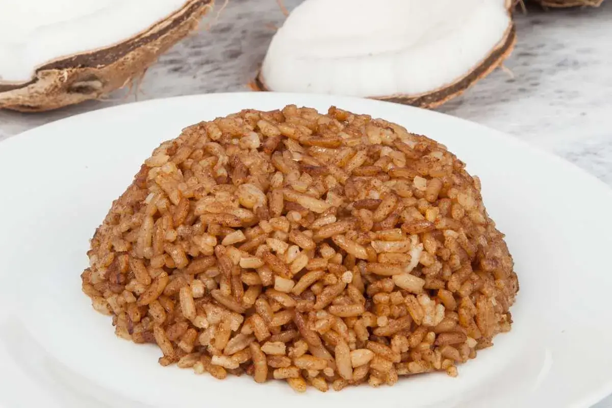 Arroz de coco servido en un plato blanco, con mitades de coco fresco al fondo. Un plato delicioso y arom&aacute;tico.