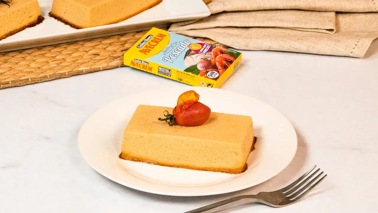 Pastel de merluza servido en plato blanco, decorado con tomate y hierbas. Al fondo, una caja de caldo de pescado Avecrem.