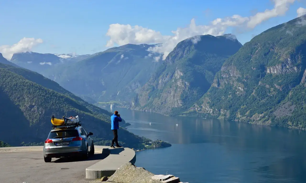 En bil med kajaker på taket parkerad vid en fjord i Norge. En man tar bilder av det dramatiska landskapet.