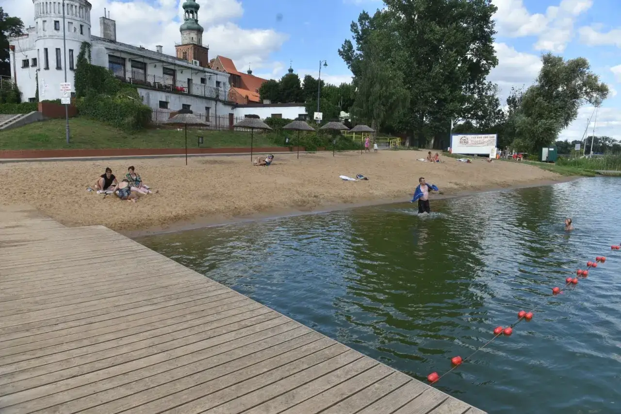 Ludzie odpoczywają na piaszczystej plaży nad jeziorem chełmżyńskim. Woda jest spokojna, a na brzegu widać drewniany pomost.