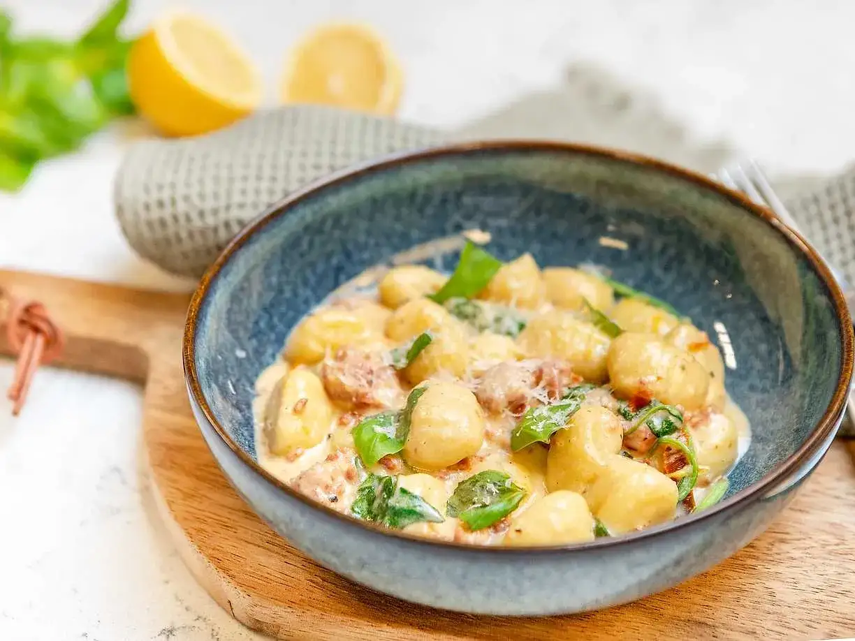 En sk&aring;l med kr&auml;mig gnocchi salsiccia, toppad med f&auml;rsk basilika och riven parmesan. Citronklyftor och en gr&aring; handduk i bakgrunden.