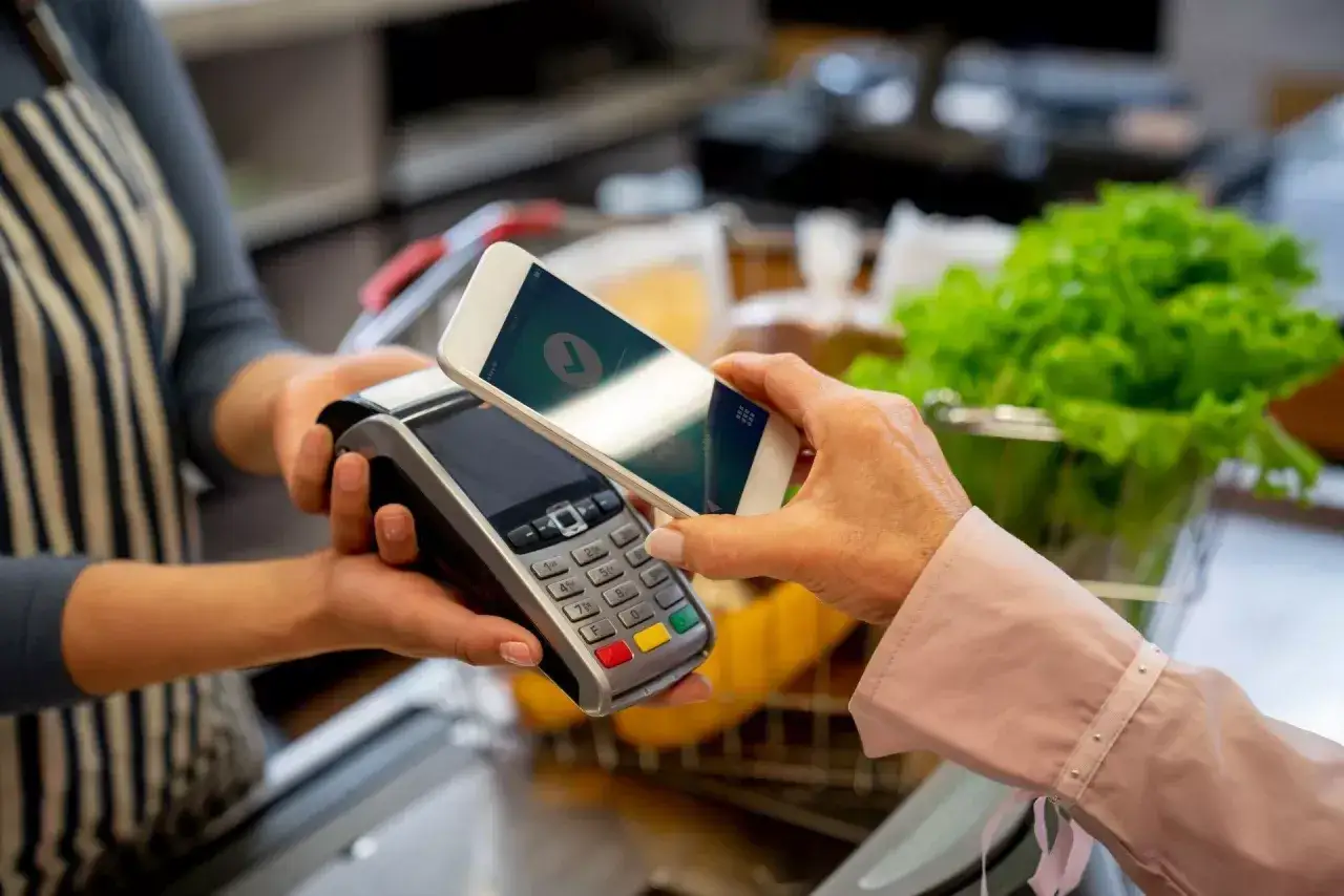 Klient płaci telefonem za zakupy, korzystając z m commerce. Na ekranie smartfona widoczny jest symbol potwierdzenia transakcji.