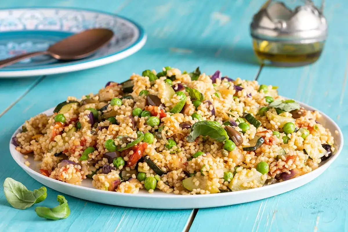 Un saporito cous cous tajine con verdure miste, piselli e basilico fresco, servito su un piatto bianco.