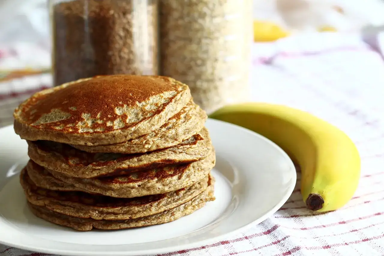 Pila di pancake senza farina, dorati e soffici, accanto a una banana matura. Sullo sfondo, barattoli di ingredienti per una colazione sana.