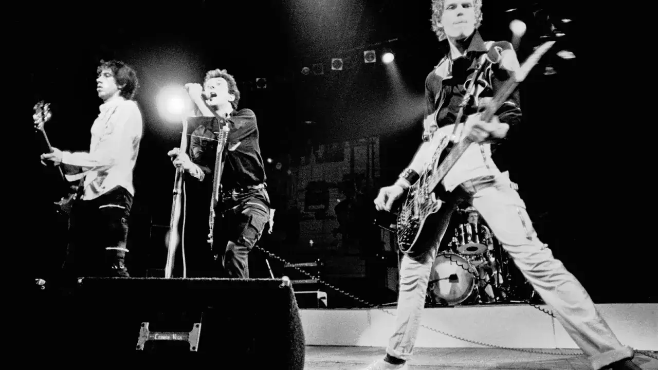 Le chanteur de The Clash, Joe Strummer, hurle au micro, entouré de ses musiciens sur scène.
