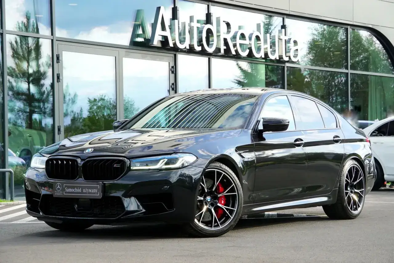 Czarny BMW M5 Competition lśni przed salonem AutoReduta. Sportowe felgi i czerwone zaciski hamulcowe podkreślają jego moc.