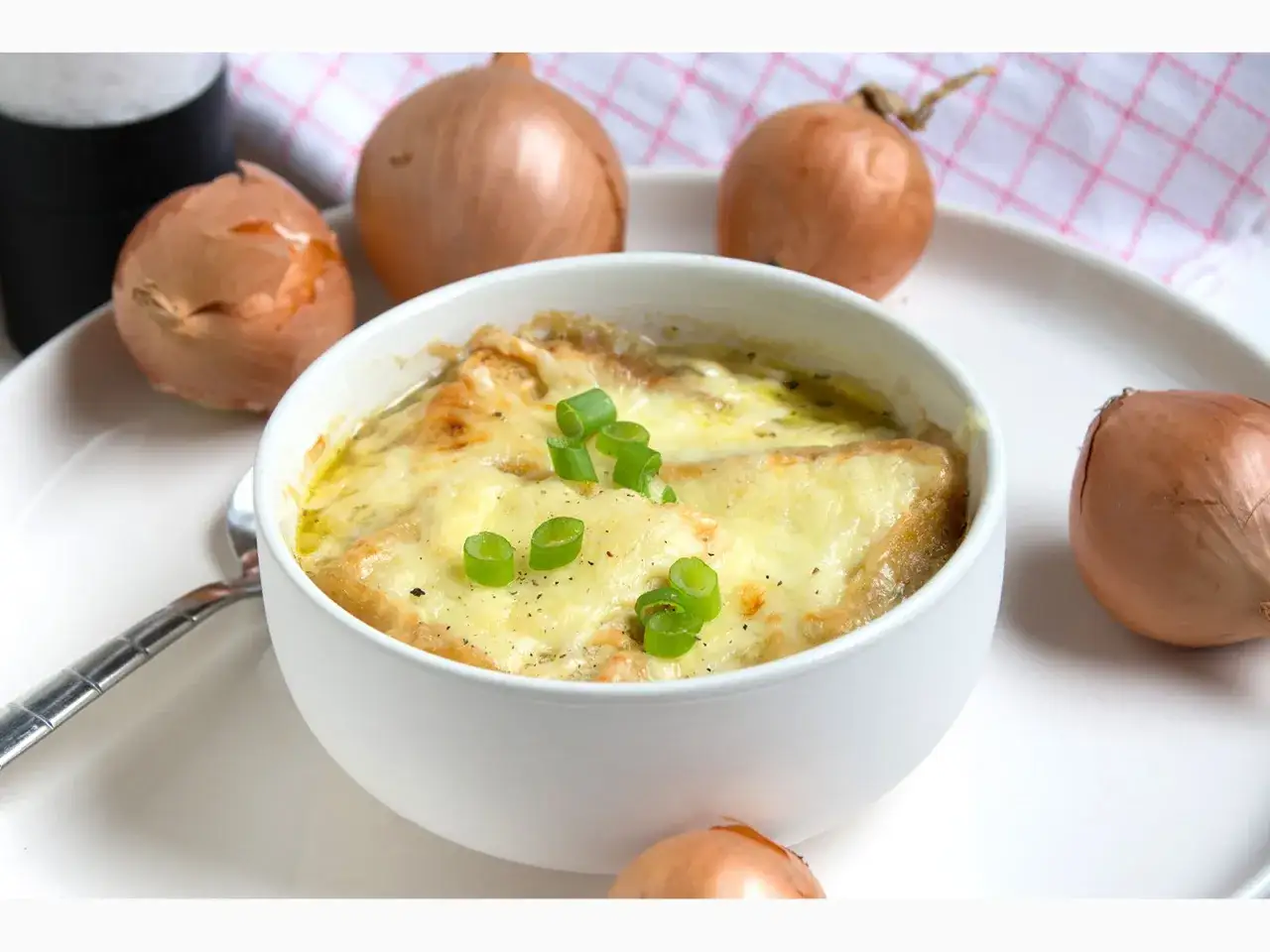 Herzhaftes Zwiebelsuppe Rezept: Eine Schüssel dampfender Zwiebelsuppe mit geschmolzenem Käse und frischen Frühlingszwiebeln, umgeben von ganzen Zwiebeln.