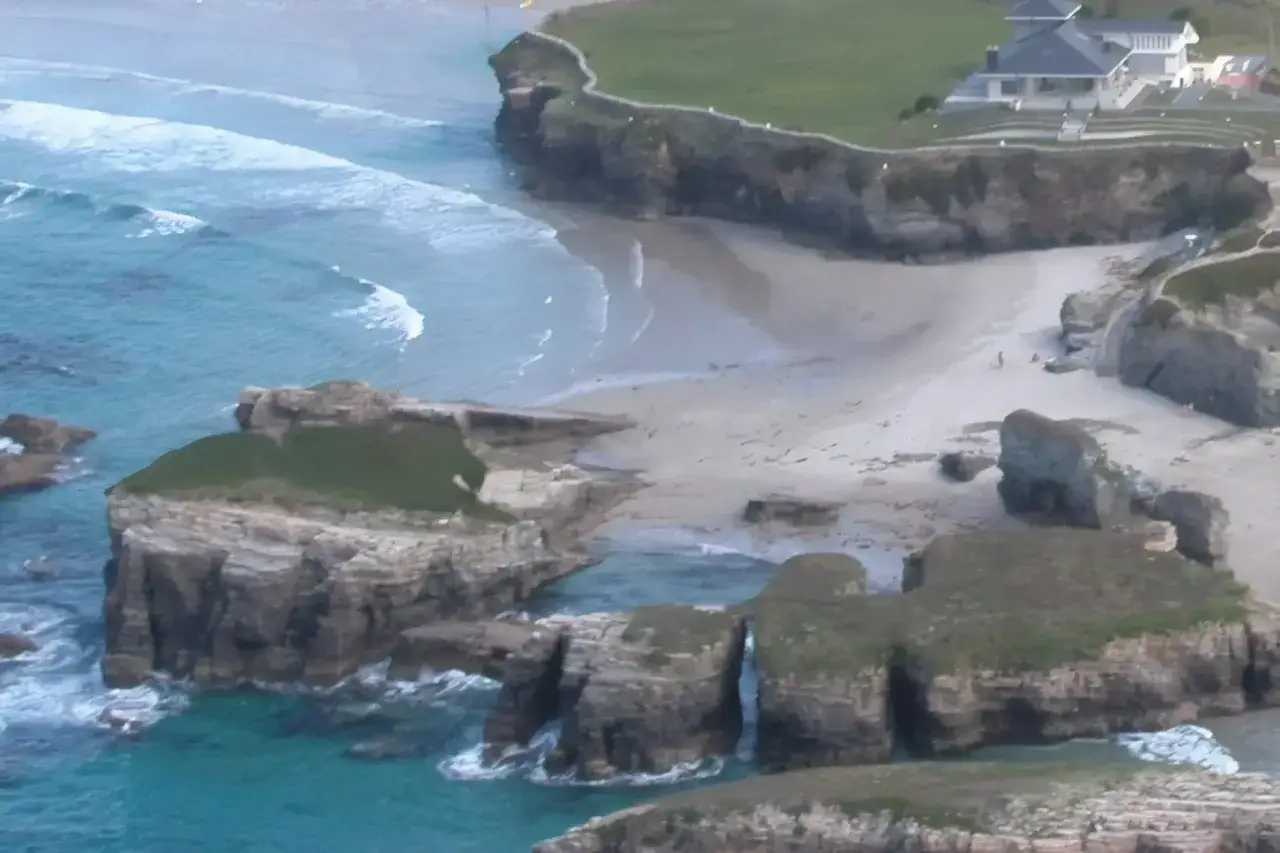 Acantilados verdes y rocosos bordean la **playa Os Castros**, donde las olas turquesas rompen suavemente en la arena dorada.