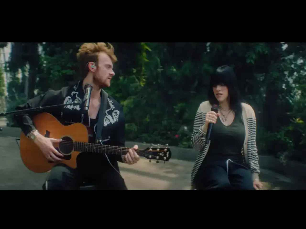 Finneas gra na gitarze, a Billie Eilish śpiewa, tworząc wzruszającą balladę w otoczeniu zieleni.