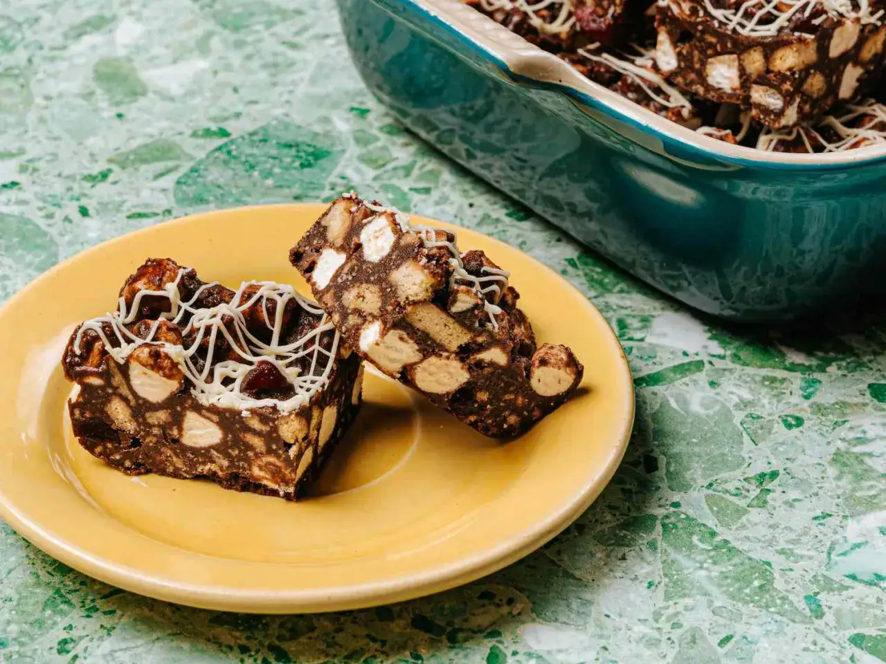 Tv&aring; bitar klassisk rocky road p&aring; en gul tallrik, med en ugnsform full av mer godsaker i bakgrunden.