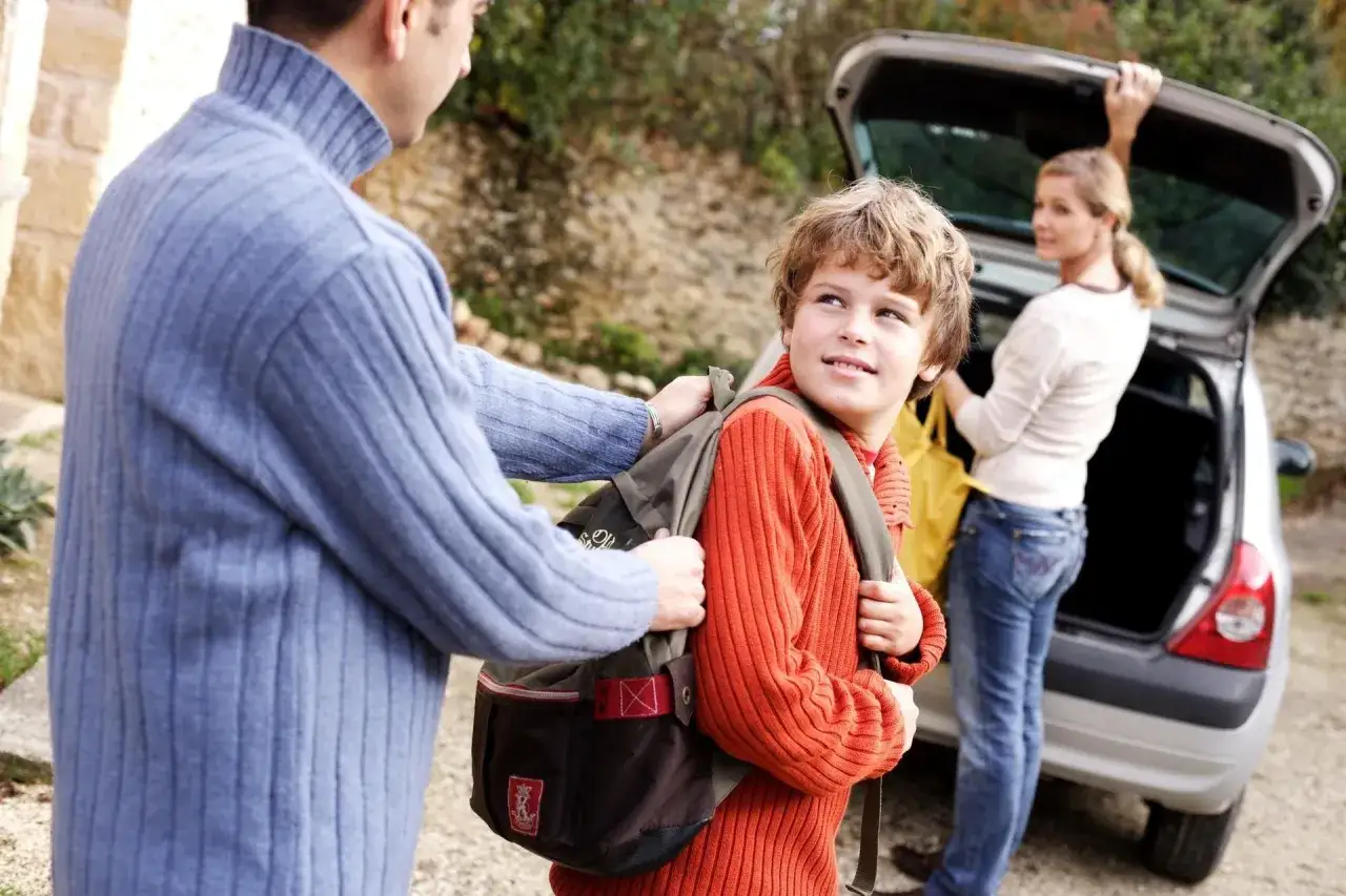 Vater hilft Sohn mit Rucksack, Mutter belädt Auto. Ein Beispiel für die praktische Umsetzung der gesetzlichen Regelung Kinderbetreuung Trennung.