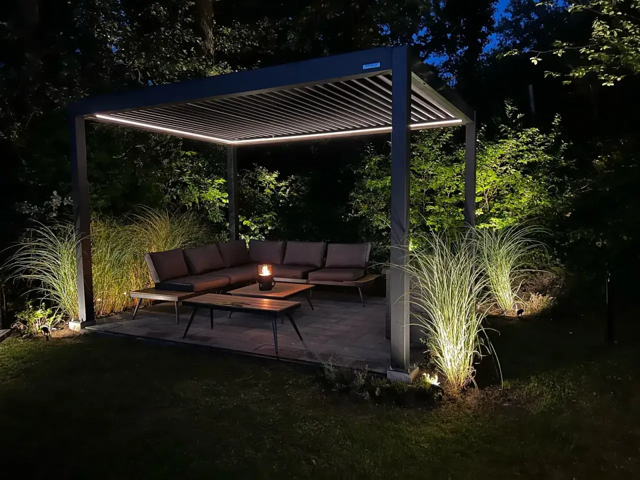 Une pergola moderne illuminée la nuit, avec un salon de jardin confortable et des herbes hautes éclairées. Une définition parfaite d'un espace extérieur invitant.