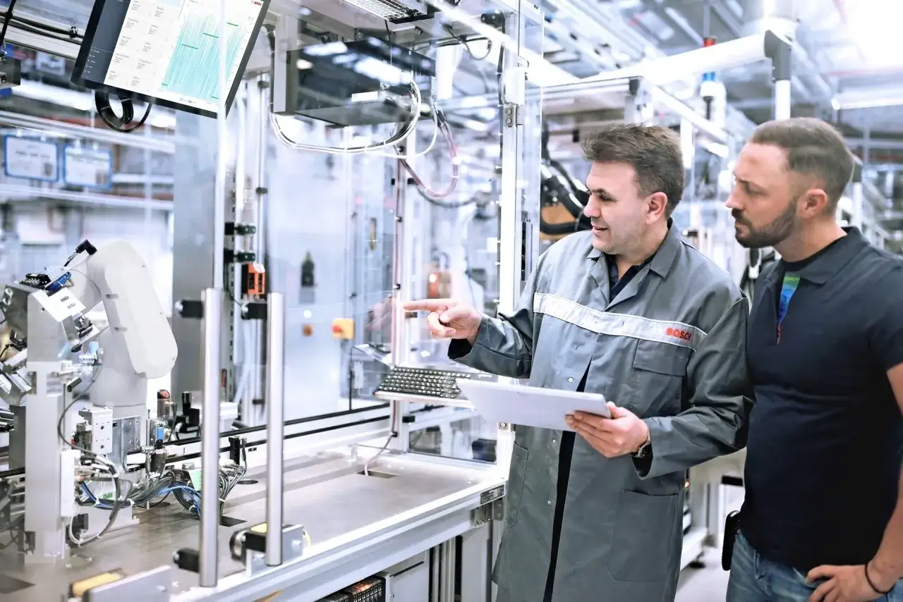 Zwei M&auml;nner besprechen eine Anlage mit einem Roboterarm. Ein Bildschirm zeigt Daten, die auf Bosch KI hindeuten.