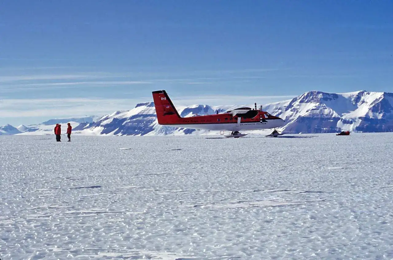 Samolot US Antarctic Program na lodzie, w tle ośnieżone góry. Dwie osoby w czerwonych kurtkach czekają na loty na Antarktydę.