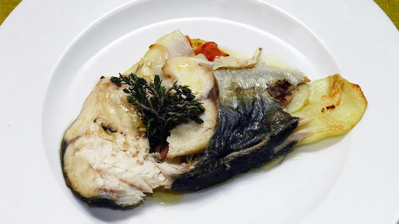 Un trozo de jurel pescado, cocinado con patatas y hierbas, servido en un plato blanco.