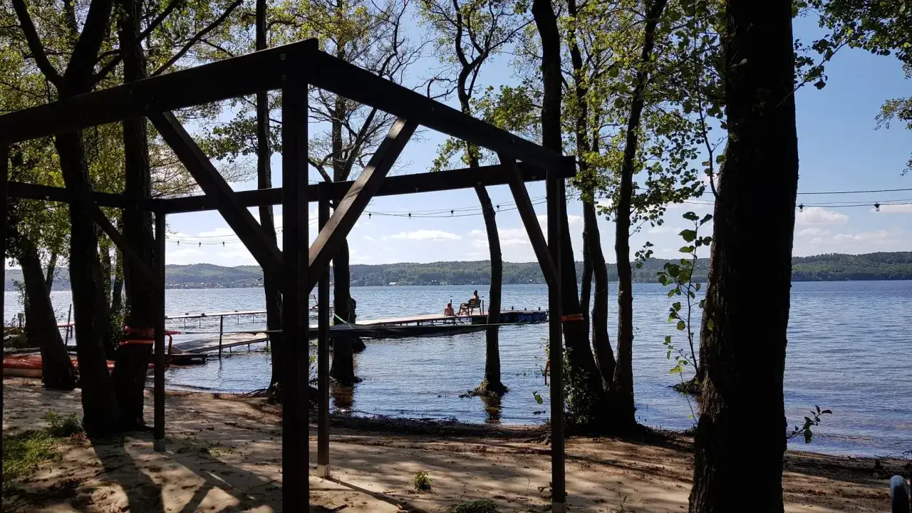 Drewniana konstrukcja na plaży, z widokiem na jezioro i pomost, gdzie odpoczywają ludzie. Idealne miejsce na relaks i odkrywanie **Władysławowo atrakcje**.