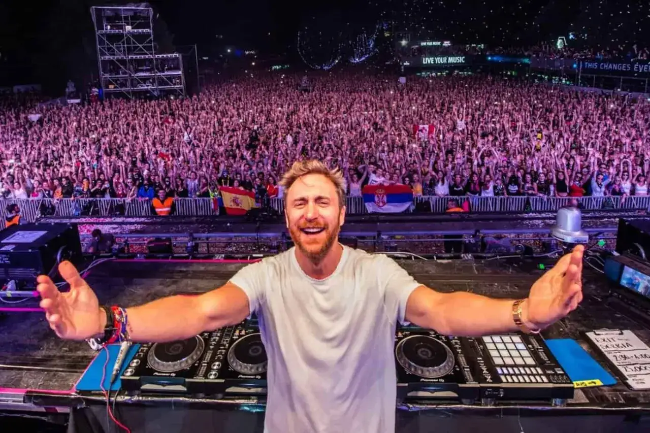 David Guetta in Italia, DJ sorridente sul palco davanti a una folla immensa, con le bandiere di Spagna e Serbia.