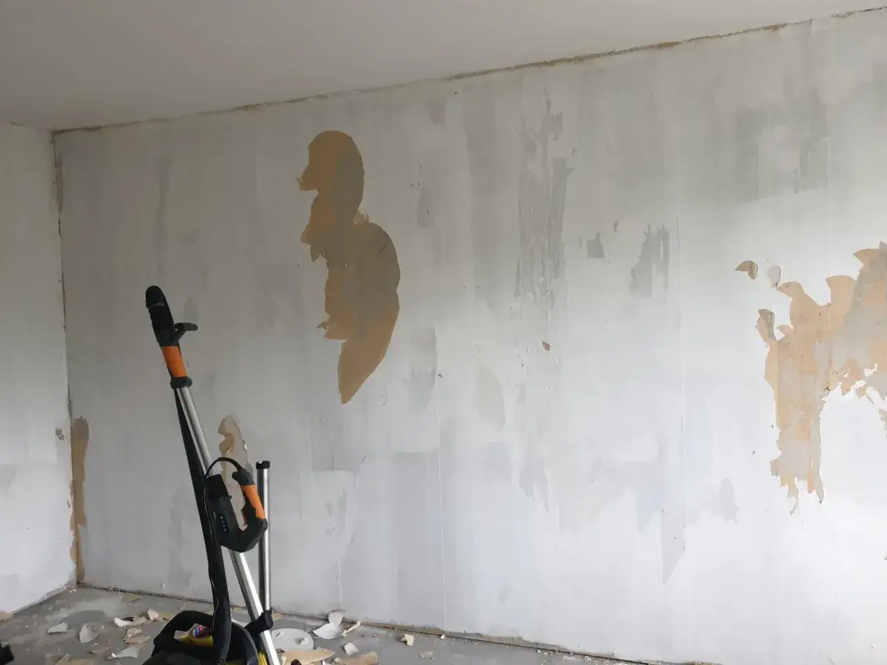 En vägg med gammal tapet som håller på att tas bort, med spår av bredspackla på tapet. En slipmaskin står redo för att jämna till ytan.