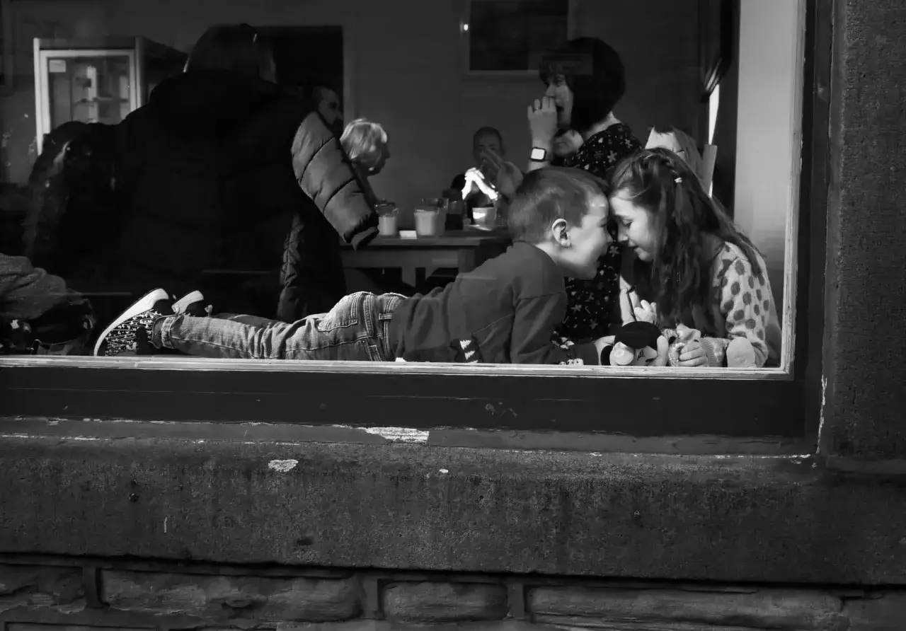 Dzieci przy oknie, jak z filmów o fotografii, dzielą się sekretami. W tle widać ludzi i stolik.