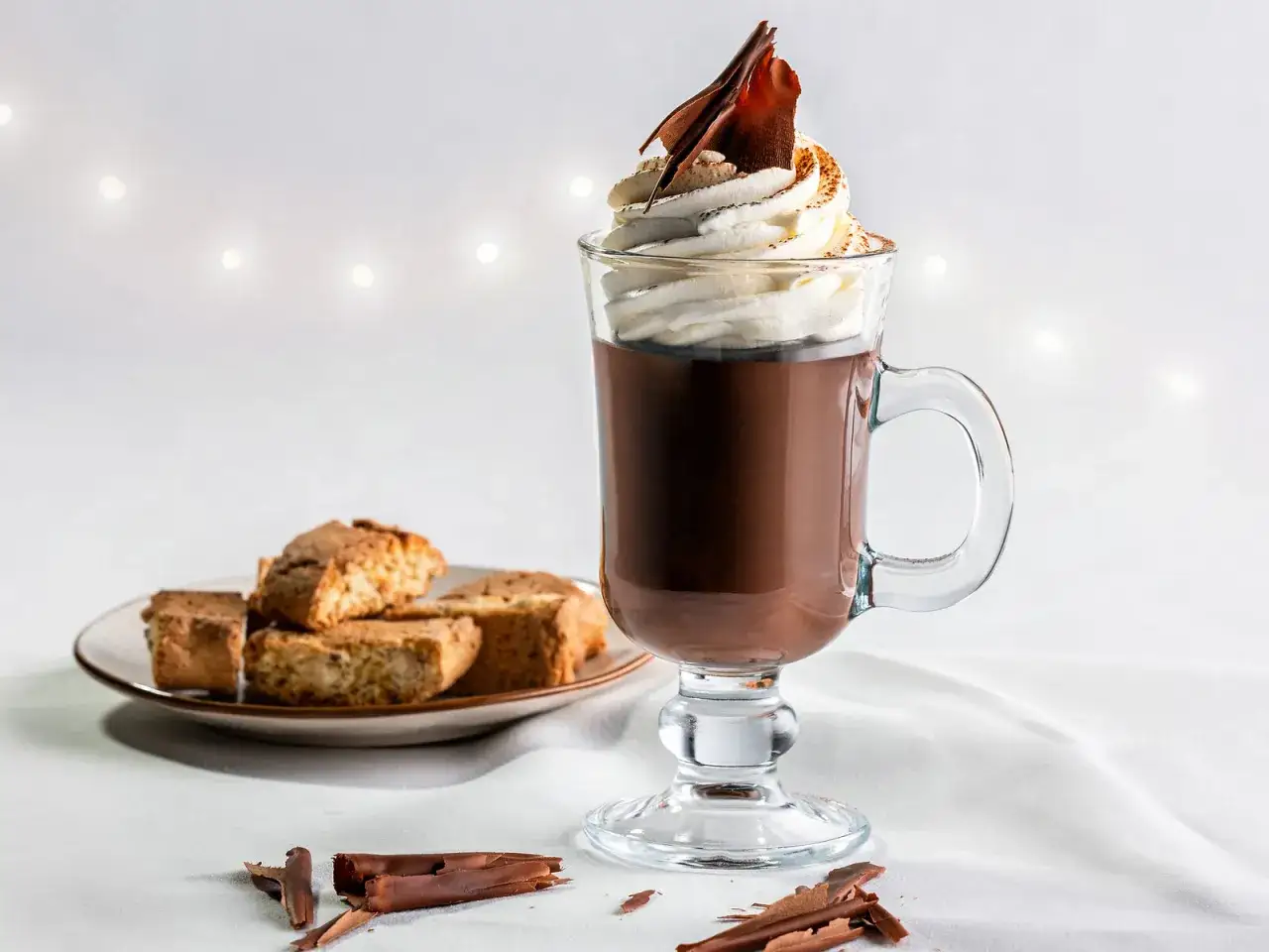 Ein Glas mit cremiger cioccolata calda, garniert mit Schlagsahne und Schokost&uuml;ckchen. Daneben Biscotti auf einem Teller.