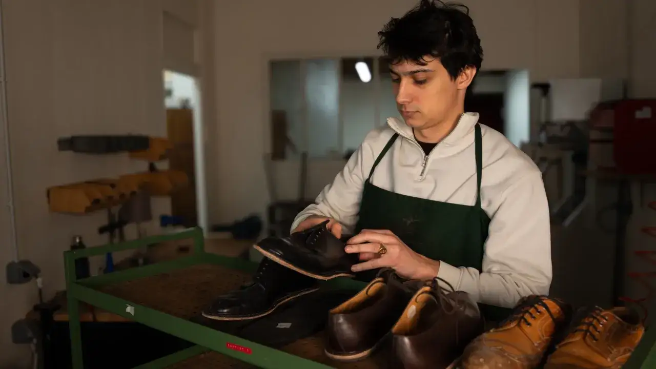 Un cordonnier examine une chaussure avant de la ressemeler. Plusieurs paires de chaussures sont dispos&eacute;es sur un &eacute;talage.
