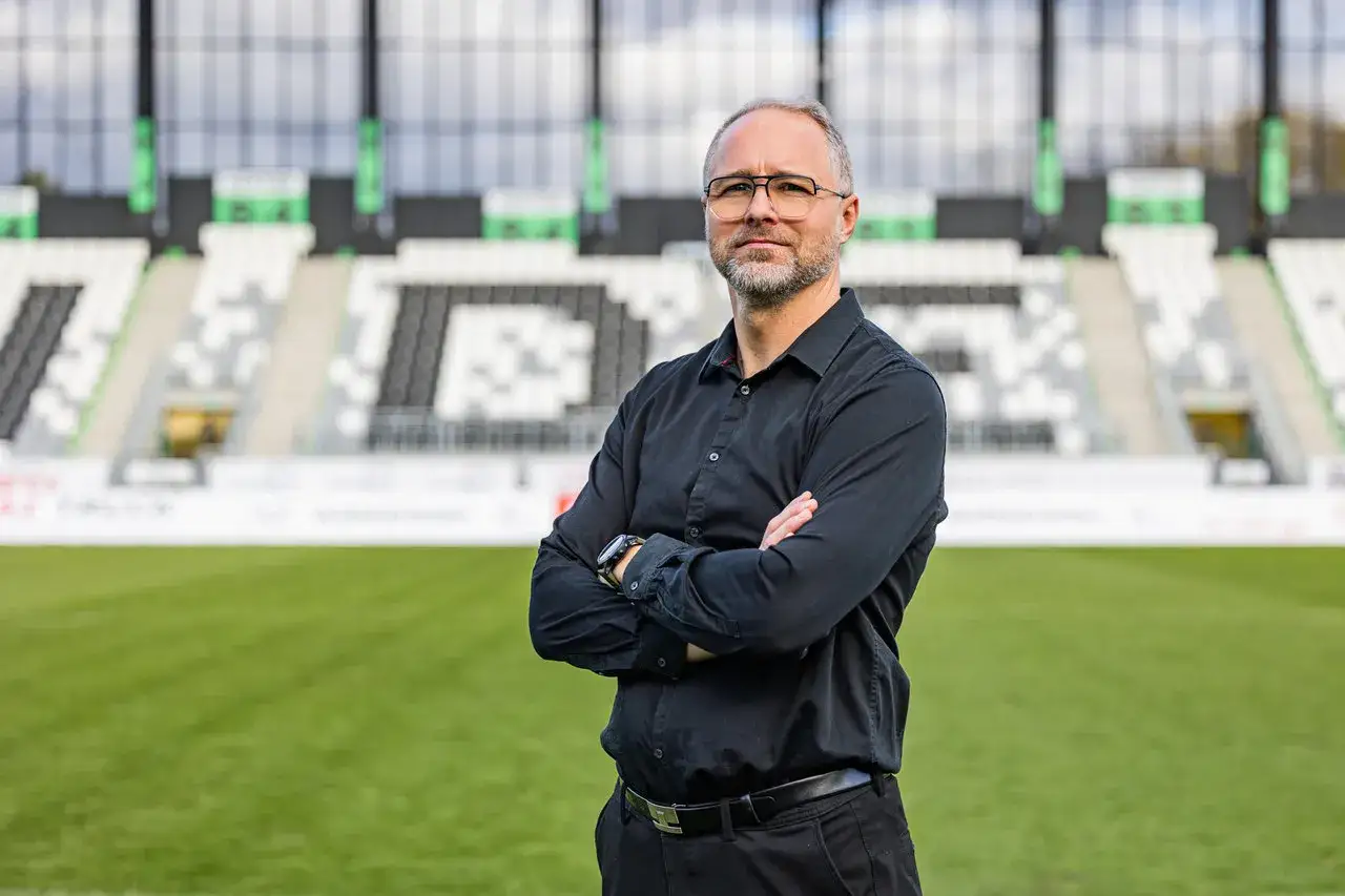 Rafał Smalec, w czarnej koszuli, stoi na tle stadionu piłkarskiego.