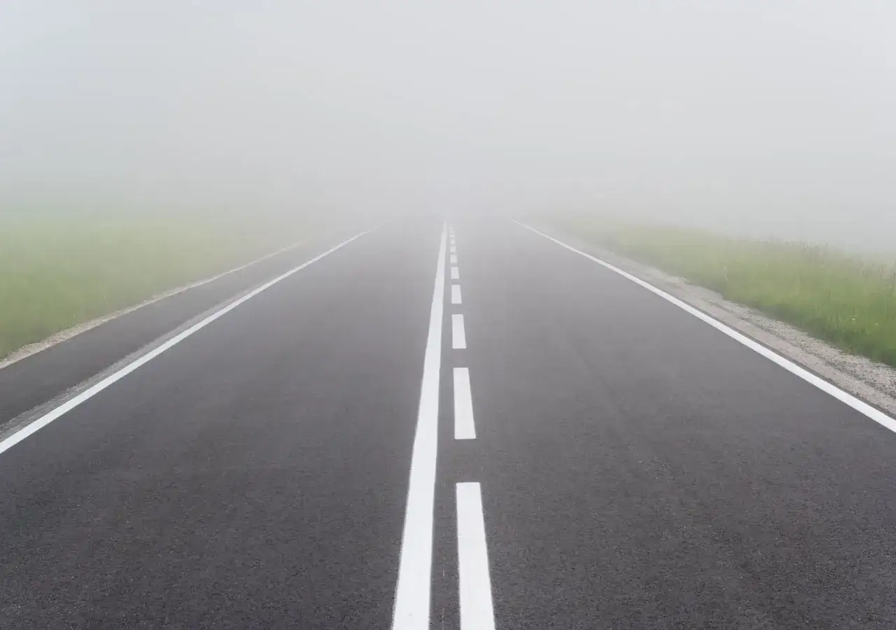 Carretera vac&iacute;a envuelta en densa niebla, creando un peligro de visibilidad. La l&iacute;nea central blanca se desvanece en la bruma.