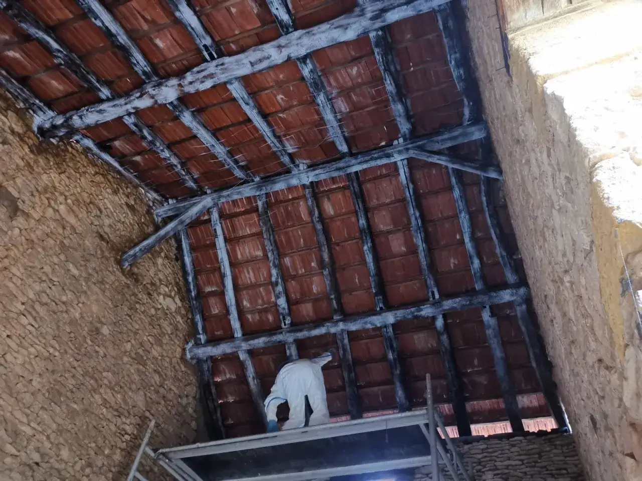Une personne en combinaison blanche travaille sur un échafaudage sous une charpente apparente bois et des tuiles rouges.