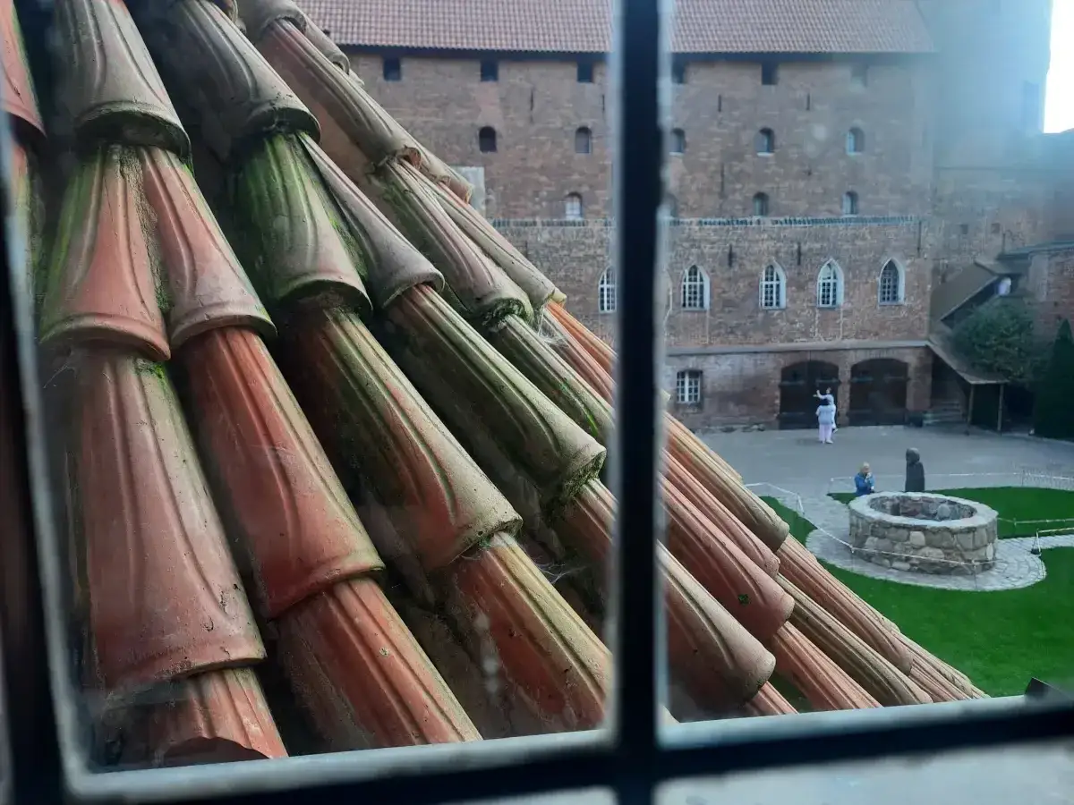 Widok z okna na dziedziniec zamku w Olsztynie. W oddali ceglane mury, studnia i spacerujący ludzie. Na pierwszym planie dachówki.