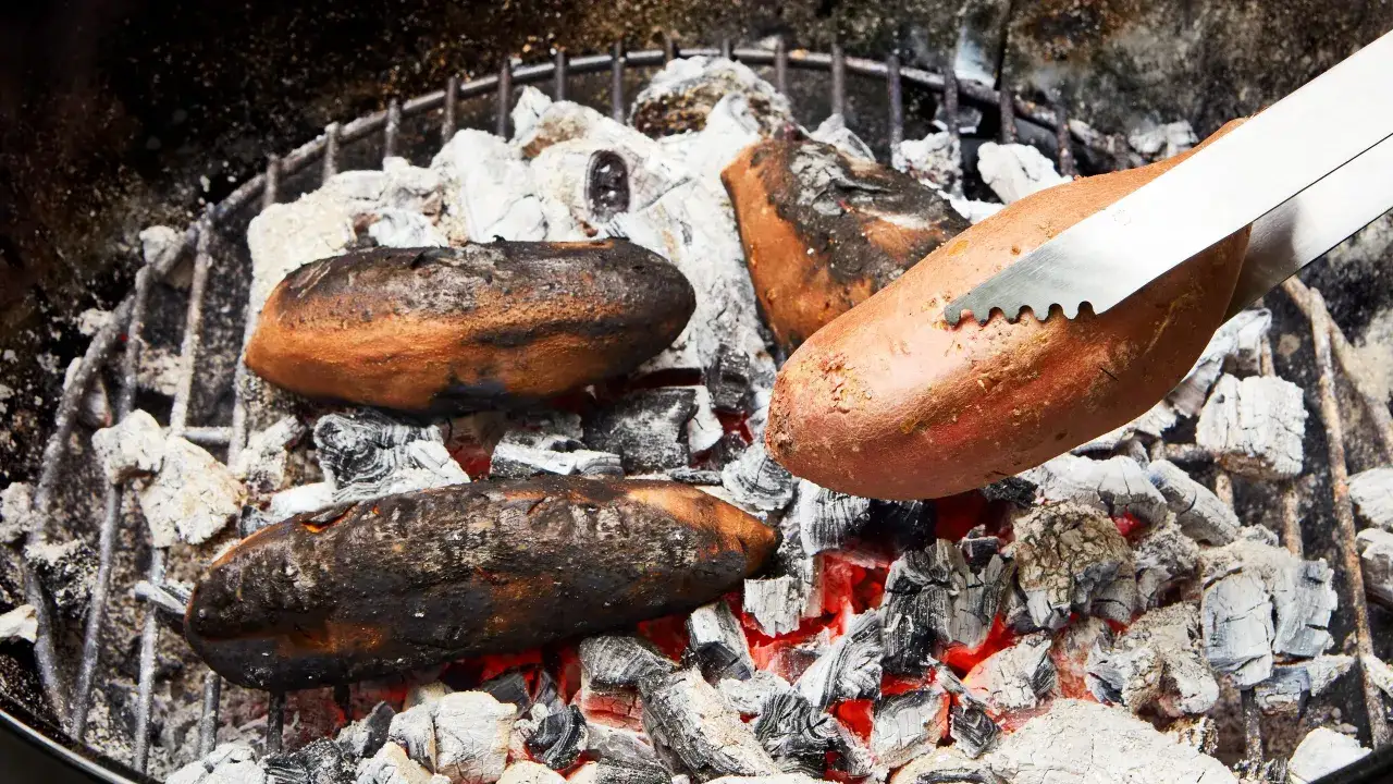 Des patates douces grillent sur des braises rouges. Un ustensile saisit une patate, montrant comment allumer un barbecue pour un repas savoureux.