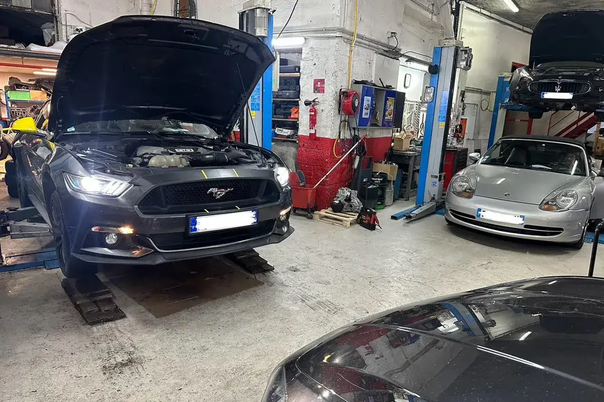 Une voiture occasion en panne après achat professionnel, une Ford Mustang, est sur un pont élévateur dans un garage. Une Porsche et une Maserati sont aussi visibles.