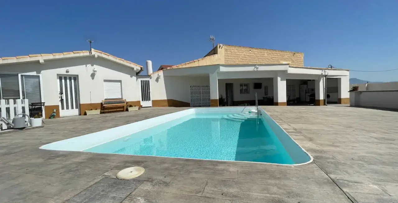 Un chalet con piscina azul cristalina y terraza de hormig&oacute;n. El sol brilla en un d&iacute;a despejado.