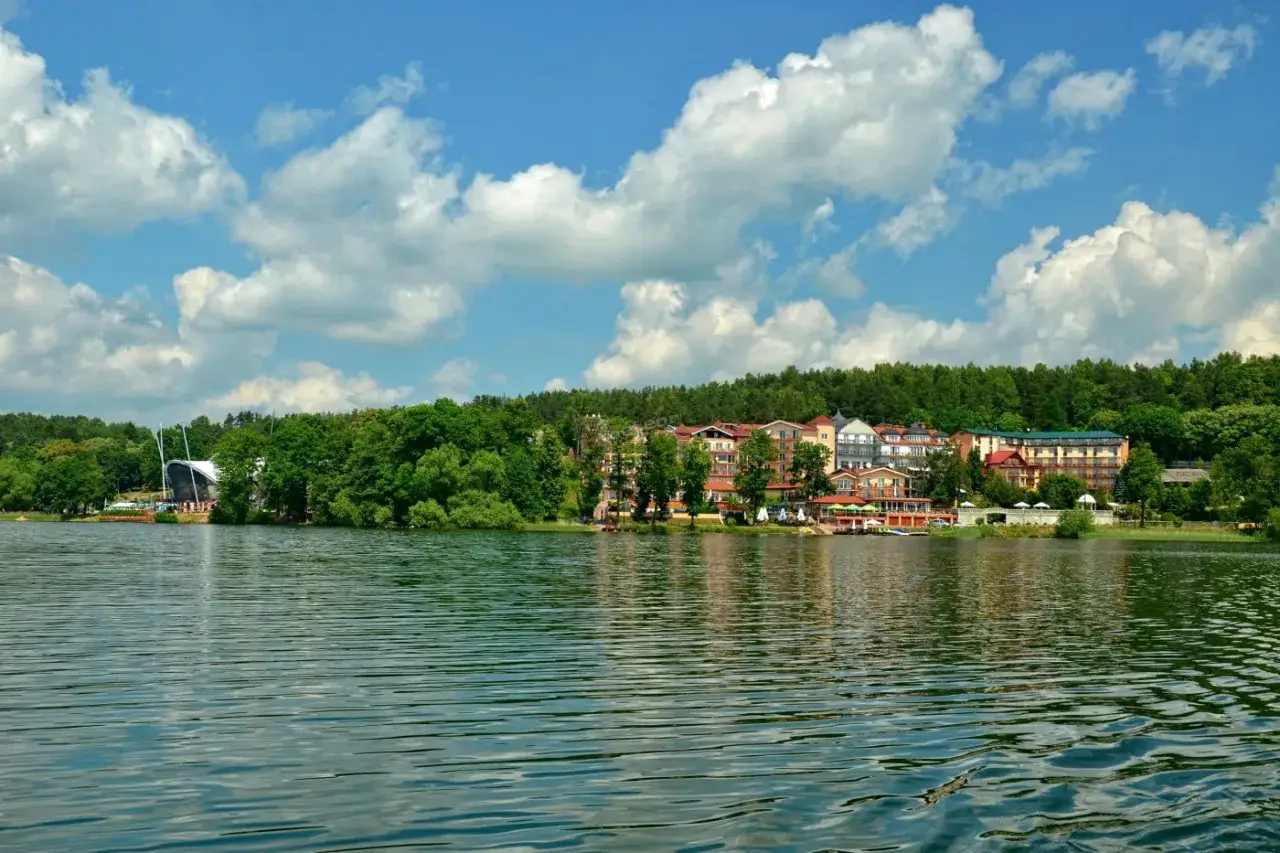 Malowniczy widok na jezioro i zabudowania w Mrągowie. W oddali widać hotel, restauracje i charakterystyczną budowlę przypominającą żagiel. To idealne miejsce na odkrywanie mrągowskich atrakcji.