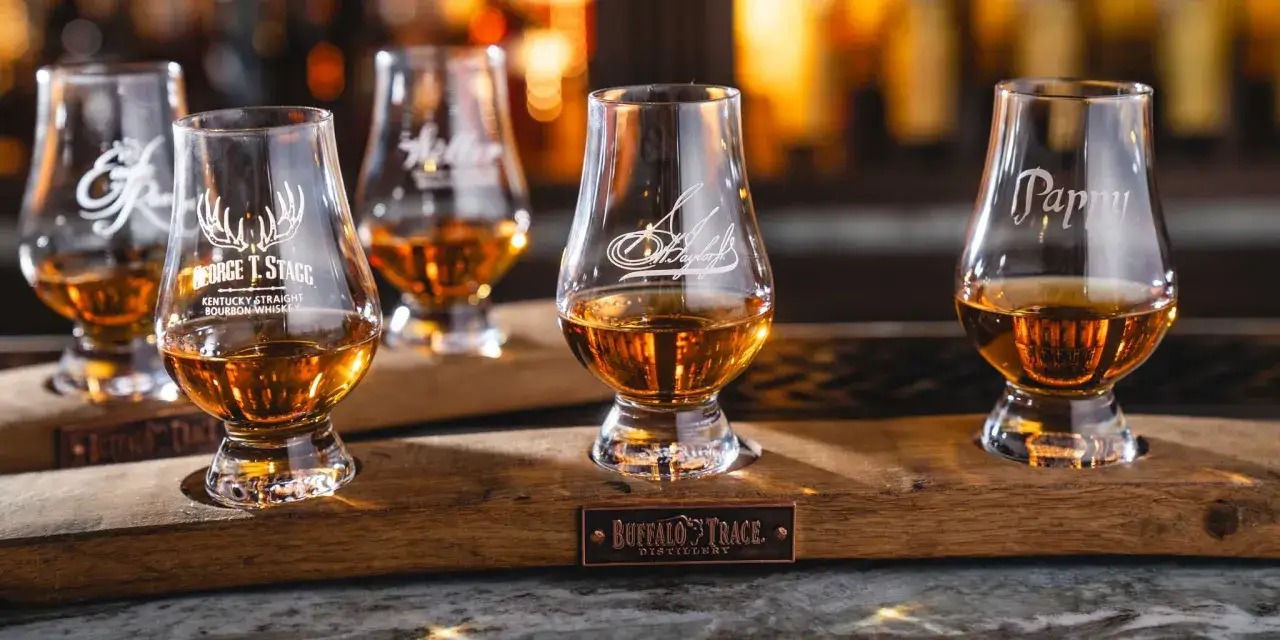 Degustacja whisky w kieliszkach z grawerem. Na drewnianej desce stoją cztery kieliszki z bursztynowym trunkiem, każdy z innym podpisem.