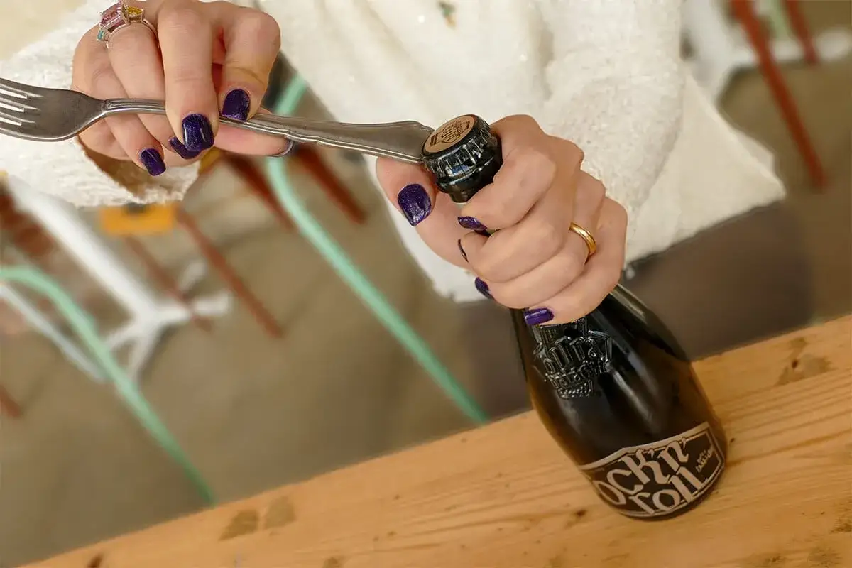 Mani con smalto viola usano una forchetta per aprire una birra.