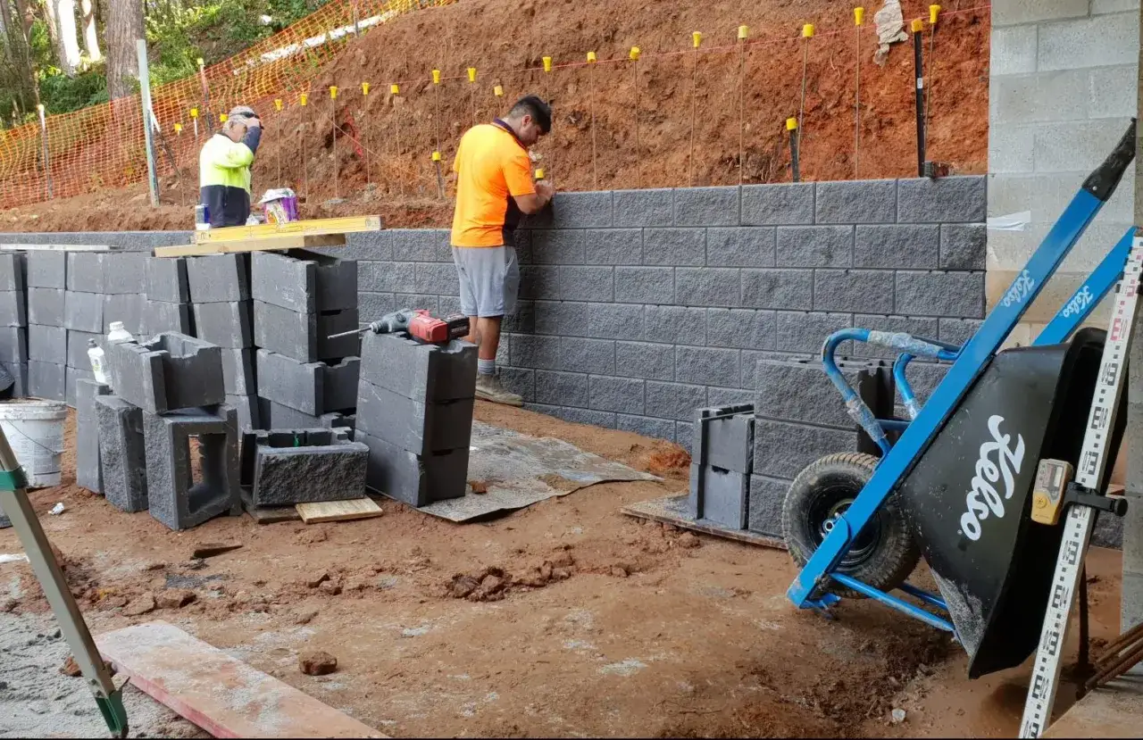 Trabajadores construyen muros de contenci&oacute;n de hormig&oacute;n. Bloques apilados y carretilla azul listos para el trabajo.