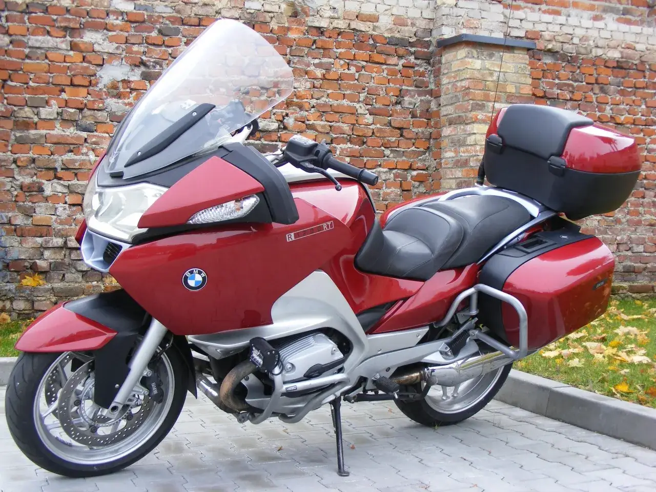 Czerwony motocykl BMW R 1200 RT z kuframi, zaparkowany przed ceglanym murem.