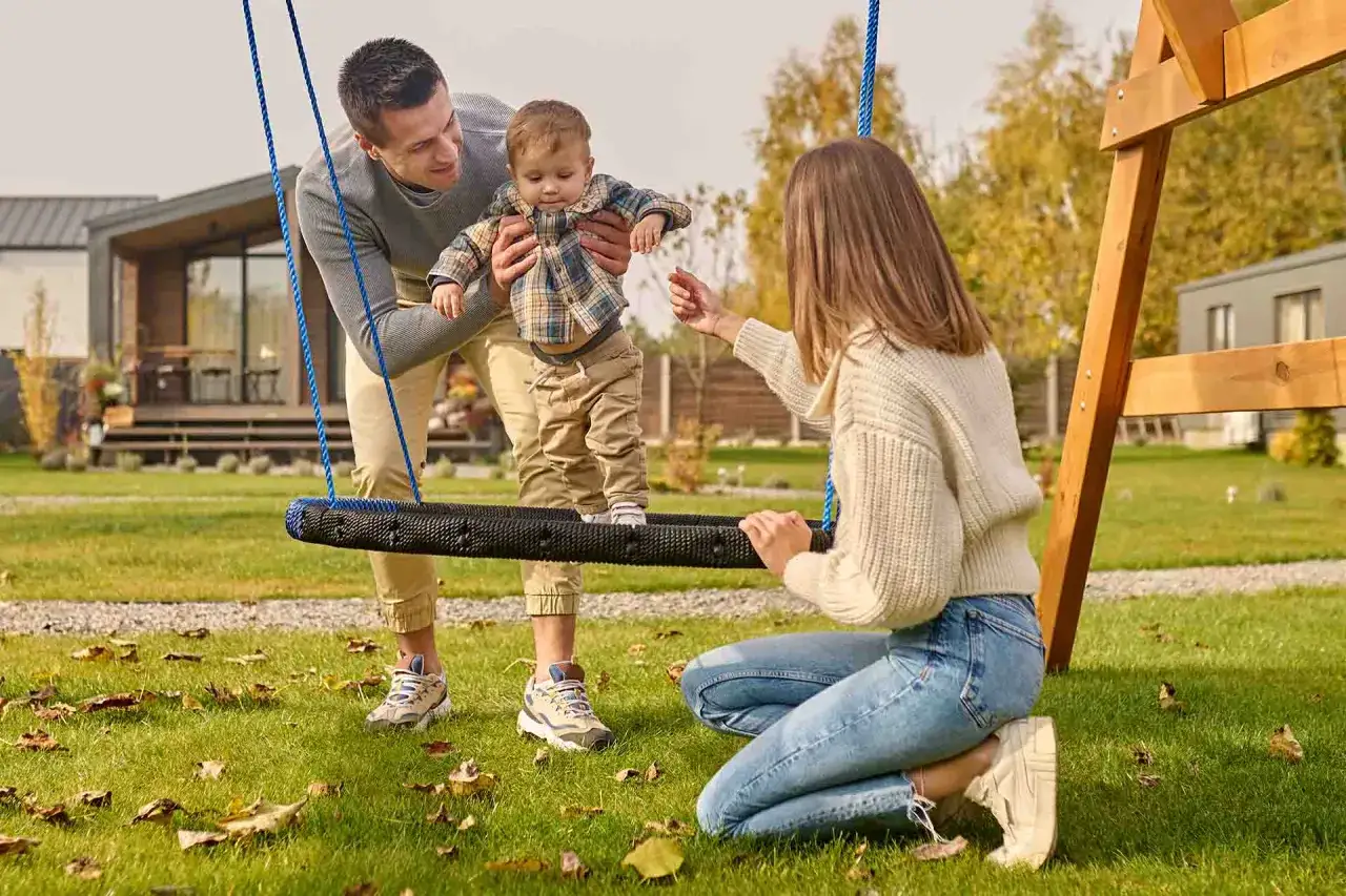 Radosne zabawy z 2 latkiem na huśtawce w ogrodzie. Tata trzyma malucha, mama zachęca do zabawy.