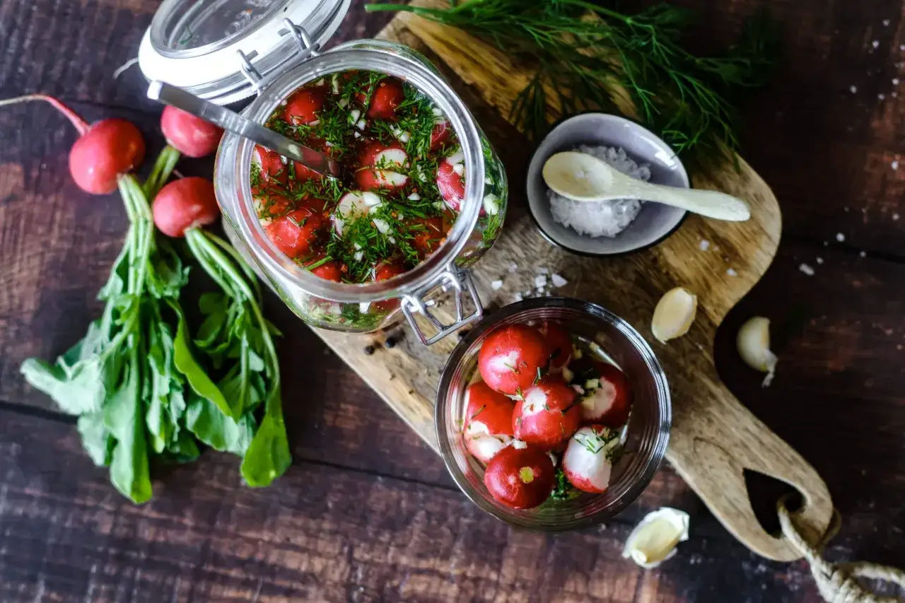 Frische Radieschen werden mit Dill und Knoblauch in einem Glasgefäß eingelegt. Ein kleiner Napf mit Salz und ein Löffel liegen daneben.