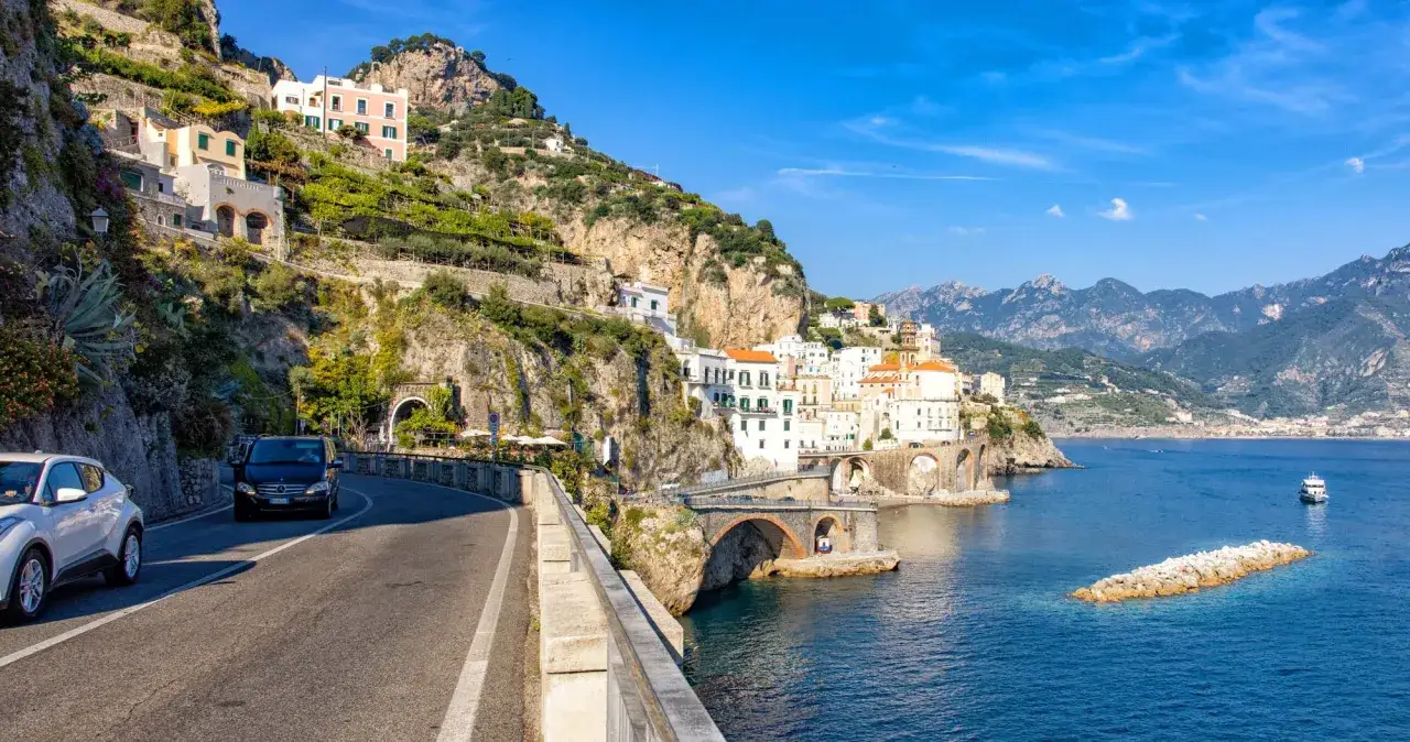 Cars drive along the scenic Amalfi road, with colorful houses clinging to the cliffs overlooking the blue sea.