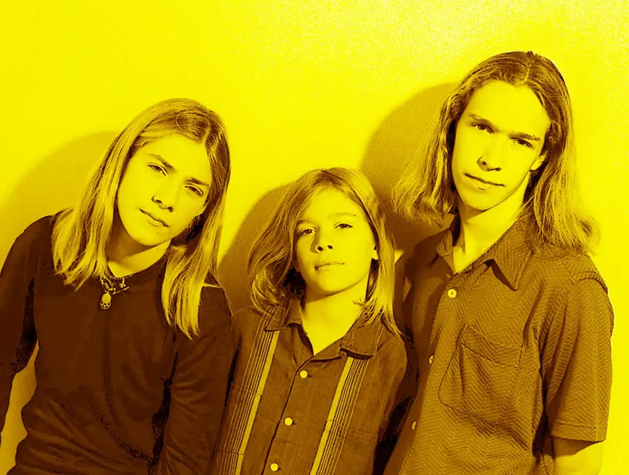 Tre unga bröder med långt blont hår, som påminner om Hanson Brothers, poserar framför en gul bakgrund.