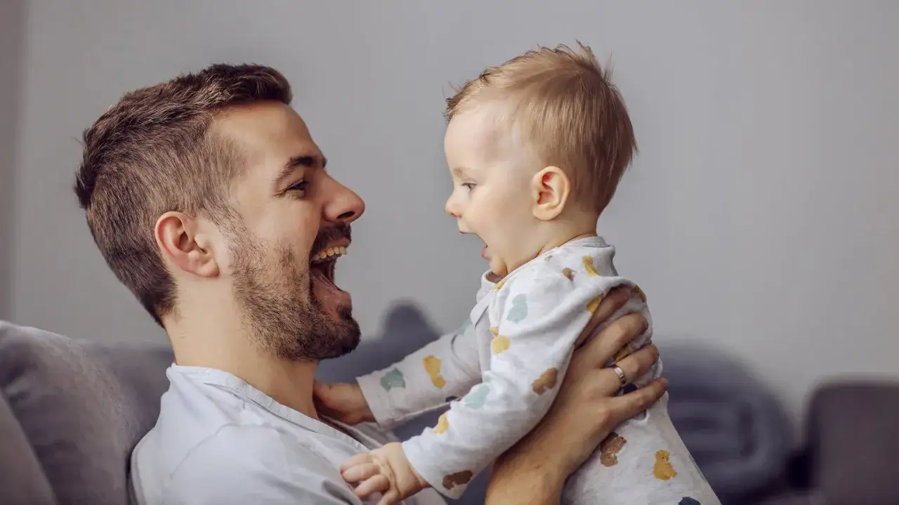 Radosny tata śmieje się do swojego 6-miesięcznego dziecka, które wyciąga do niego rączki.