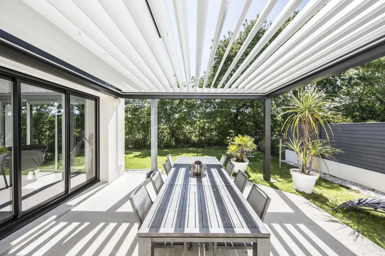 Tavolo da pranzo all'aperto sotto una pergola bioclimatica. La struttura moderna offre ombra e riparo, ideale per godersi il giardino. Informati sui permessi necessari per la tua pergola bioclimatica.