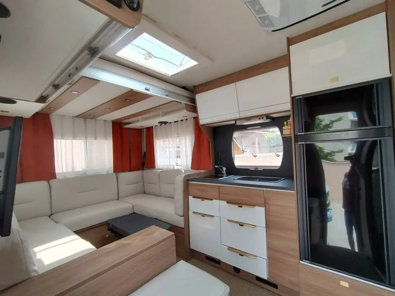 L'espace de vie arri&egrave;re d'un camping-car, avec un canap&eacute; en L blanc, une cuisine &eacute;quip&eacute;e et un r&eacute;frig&eacute;rateur.