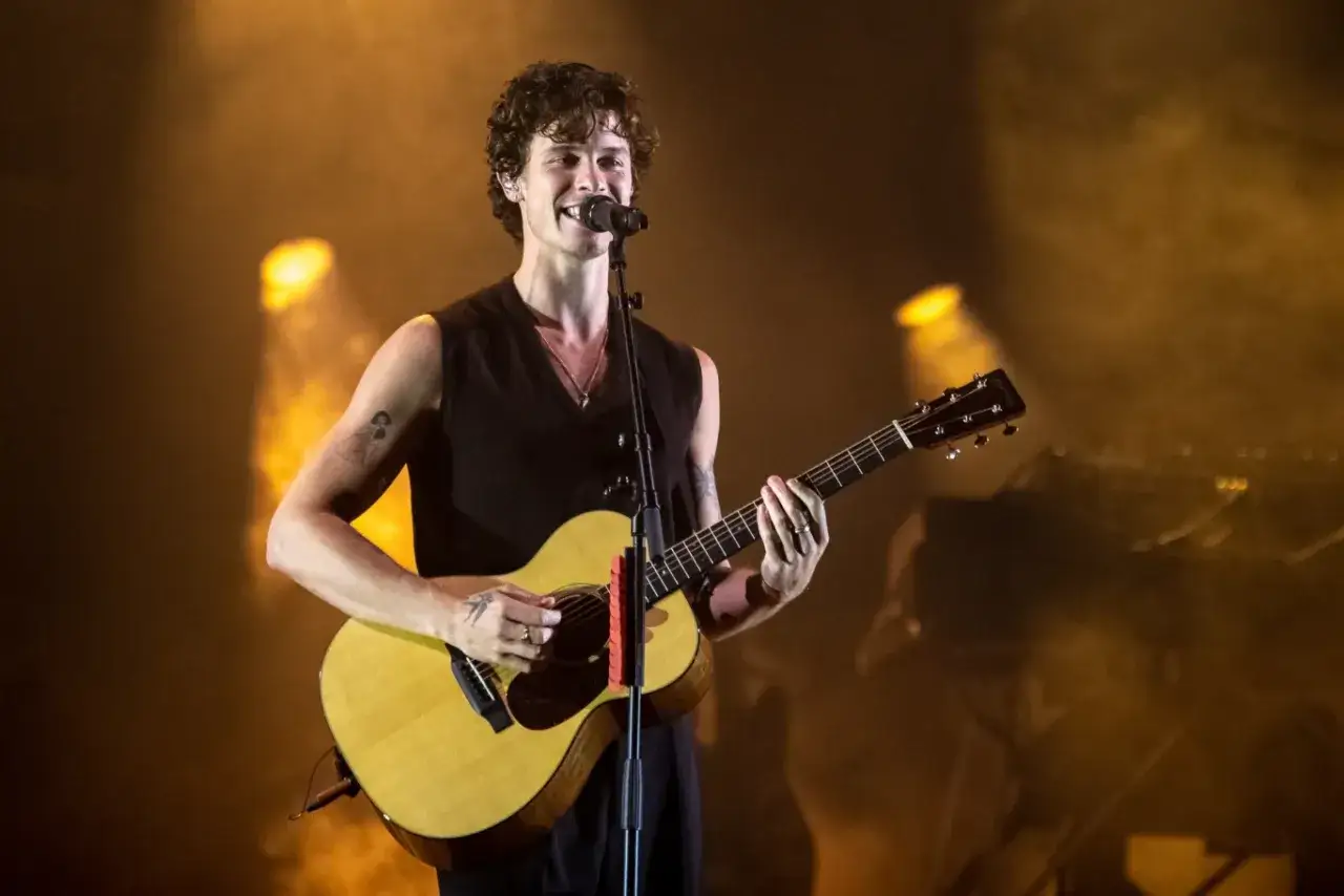 Uśmiechnięty piosenkarz Shawn Mendes gra na gitarze akustycznej na scenie, oświetlony ciepłym, pomarańczowym światłem.