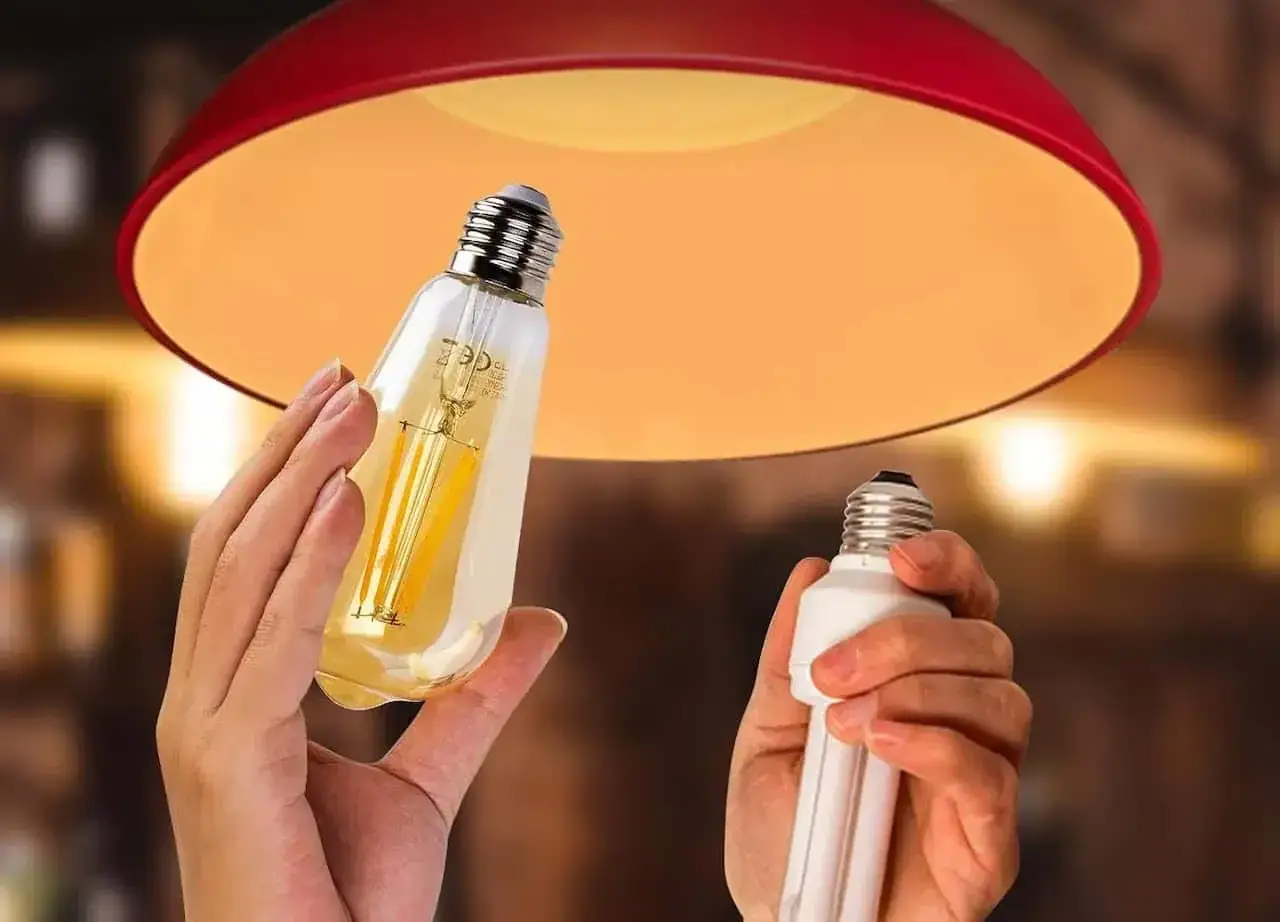 Une ampoule connectée orange, au design vintage, est tenue à côté d'une ampoule fluocompacte, sous un abat-jour rouge.