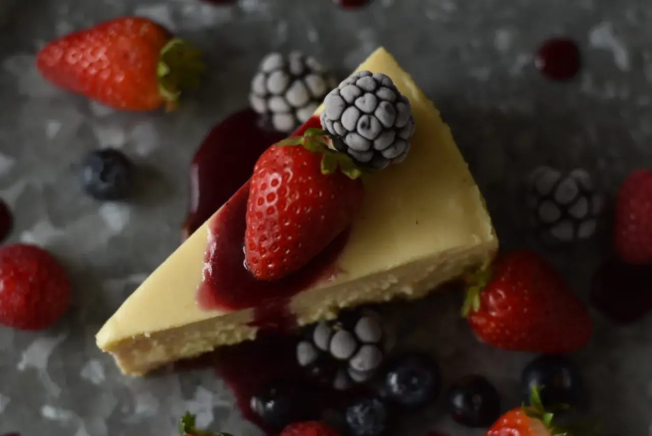 Tranche de cheesecake sans gluten sans lactose, garnie de fraises, m&ucirc;res givr&eacute;es et d'un coulis de fruits rouges.