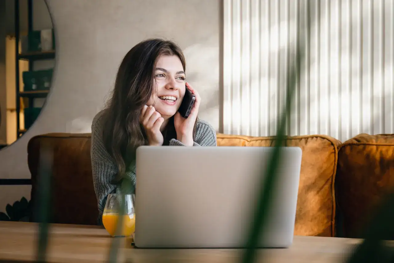 Uśmiechnięta kobieta rozmawia przez telefon, siedząc przy laptopie. Dowiaduje się, jak zarobić 100 zł dziennie przez internet.