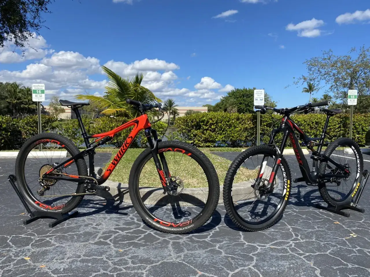 Two mountain bikes, one orange and one black, are parked on a paved surface. The orange bike is a Specialized S-Works, a type of downcountry bike designed for versatile trail riding.